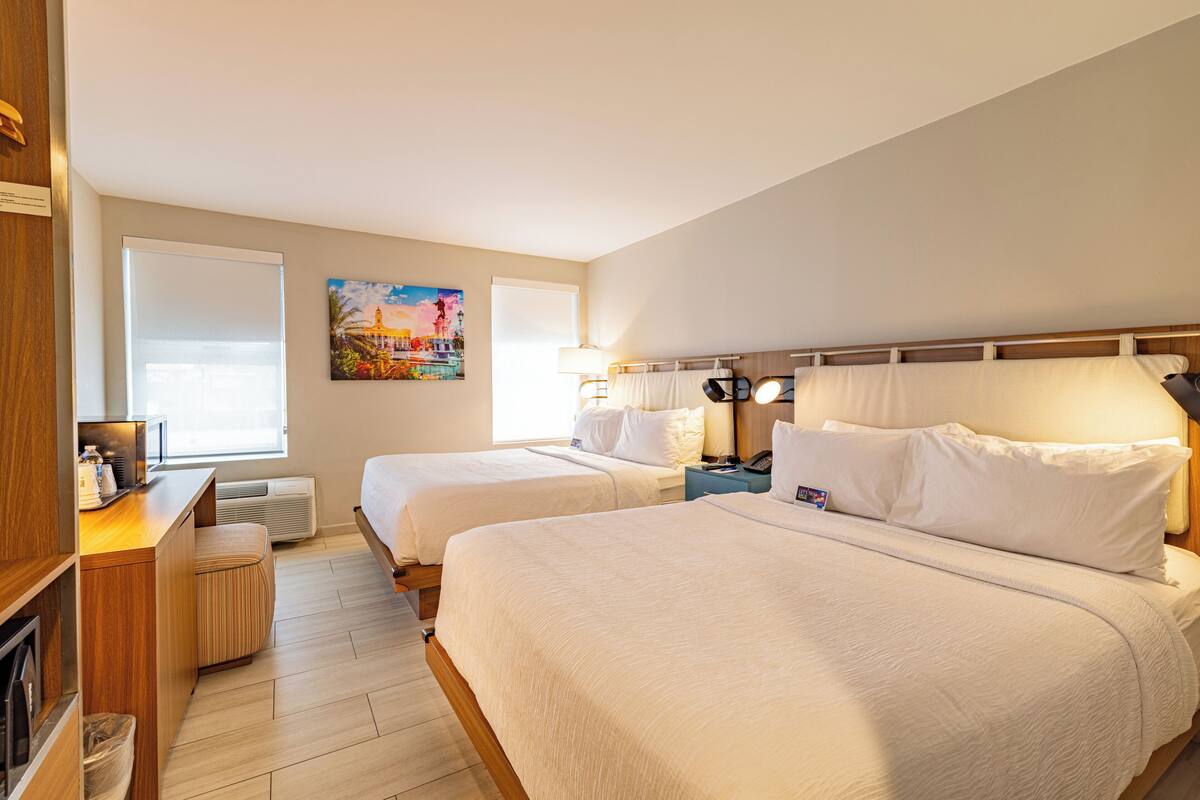 A hotel room with two beds, a desk, a lamp, and a painting on the wall.