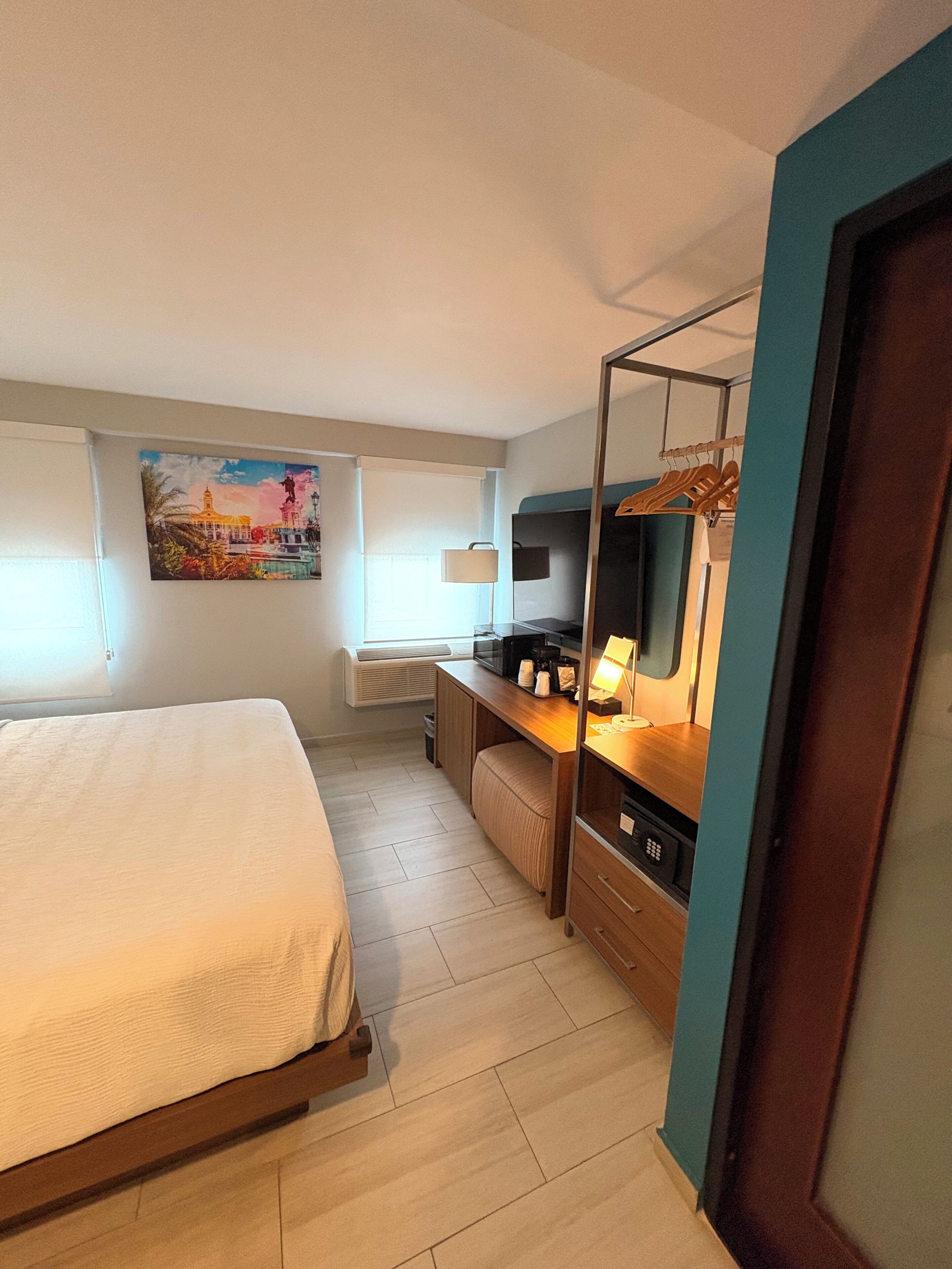 A hotel room with a bed, a desk with a computer, a TV, and a painting on the wall.