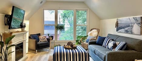 A cozy living room with a fireplace, a television, a sofa, and a large window offering a view of the outdoors.
