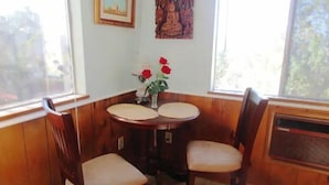 A corner room with wooden paneled walls, a small table with two chairs, a window with a view of trees, and a wall-mounted air conditioner.