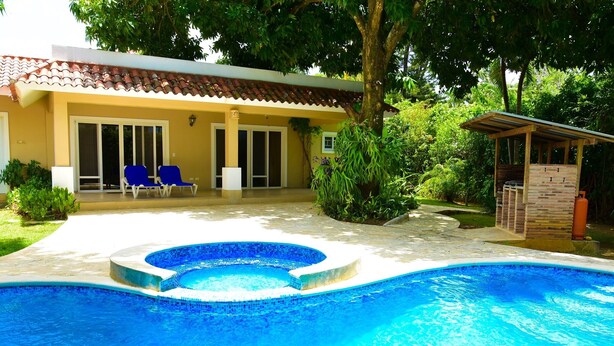Une piscine bordée de carreaux, un patio couvert avec des chaises bleues, et un bâtiment doté d’un toit en tuiles et de portes-fenêtres coulissantes.