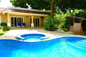 Una piscina con bordo piastrellato, un portico coperto con sedie blu e un edificio con tetto in tegole e porte scorrevoli in vetro.