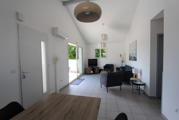 A spacious living area with a dining table, a sofa, and a TV. There is a sliding glass door leading to a balcony.