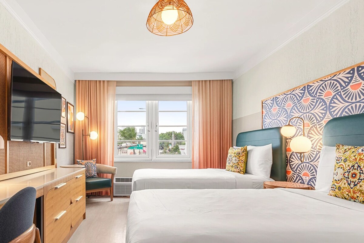 A hotel room with a bed, a desk with a TV, a chair, a window with curtains, and a patterned headboard.