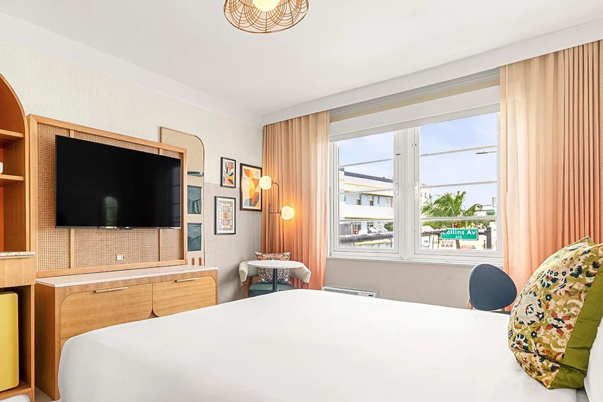 A hotel room with a bed, a television, a desk with a chair, a window with curtains, and a kitchenette area.