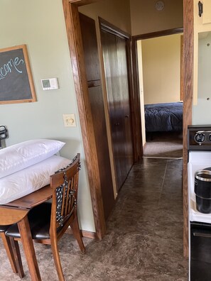 Ein Zimmer mit einem Bett, einem Holzstuhl, einem Tisch mit einem Kissen, einem Herd und einem an der Wand befestigten Thermostat.