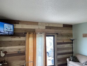 Ein Zimmer mit Holzwandverkleidung, einer Schiebetür aus Glas, einem an der Wand montierten Fernseher und einem Sofa mit Kissen.