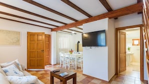 Ein Wohnzimmer mit Holzboden, einem an der Wand befestigten Fernseher, einem Essbereich mit Tisch und Stühlen sowie einer durch eine offene Tür einsehbarer Küche.