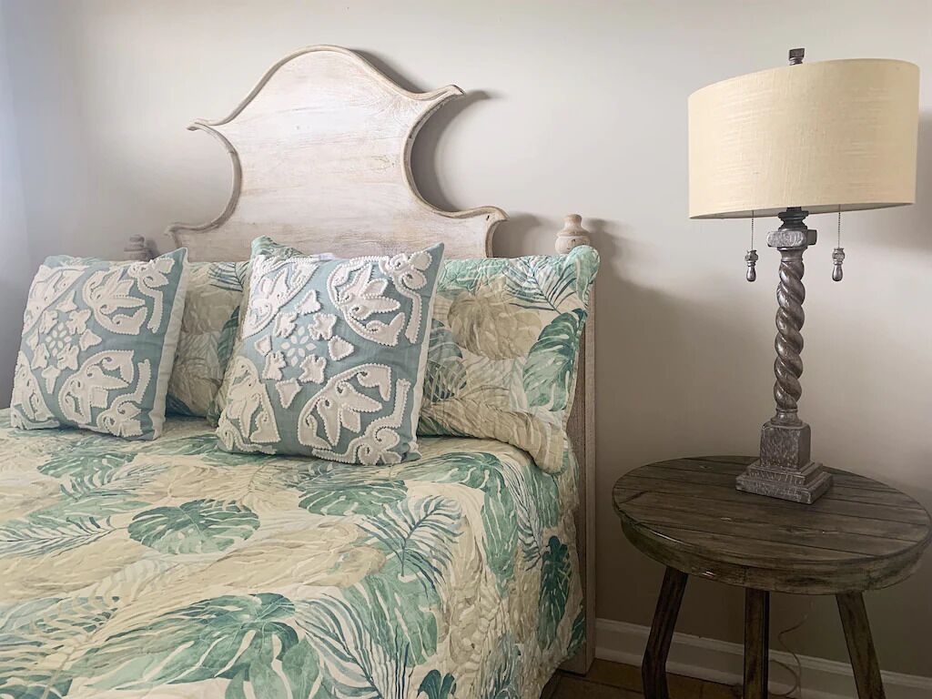 Un letto con una coperta e dei cuscini a motivi floreali, una testiera in legno e un piccolo tavolino rotondo con una lampada accanto.