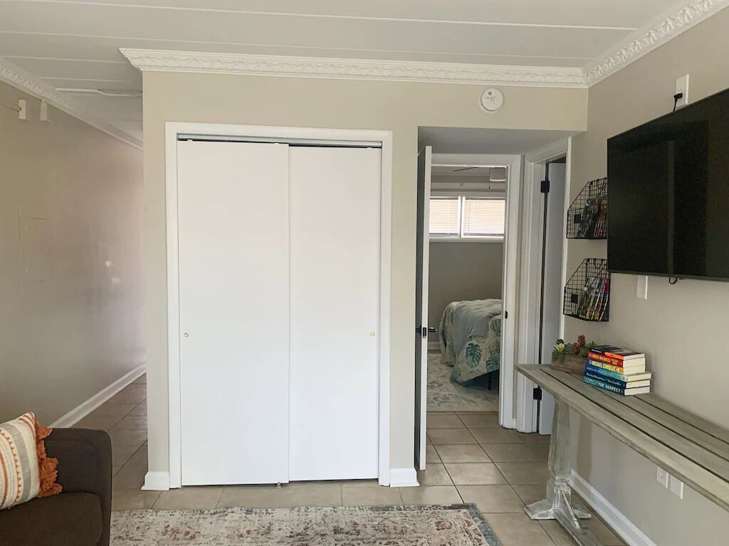 A living room with a closed white door, a sofa with a patterned throw, a TV on a stand, and a hallway leading to a bedroom.