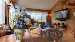 A wooden interior with a dining table, floral vase, and a television mounted on the wall. - One-of-a-kind 4 Acre Adirondack Escape Short Drive to LG & GORE Mountain!
 (Warrensburg)