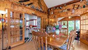 A wooden cabin interior with a dining area, kitchen, and large windows. - One-of-a-kind 4 Acre Adirondack Escape Short Drive to LG & GORE Mountain!
 (Warrensburg)