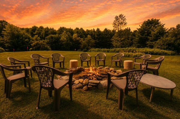 A circular arrangement of chairs around a fire pit in a grassy outdoor area with trees in the background.