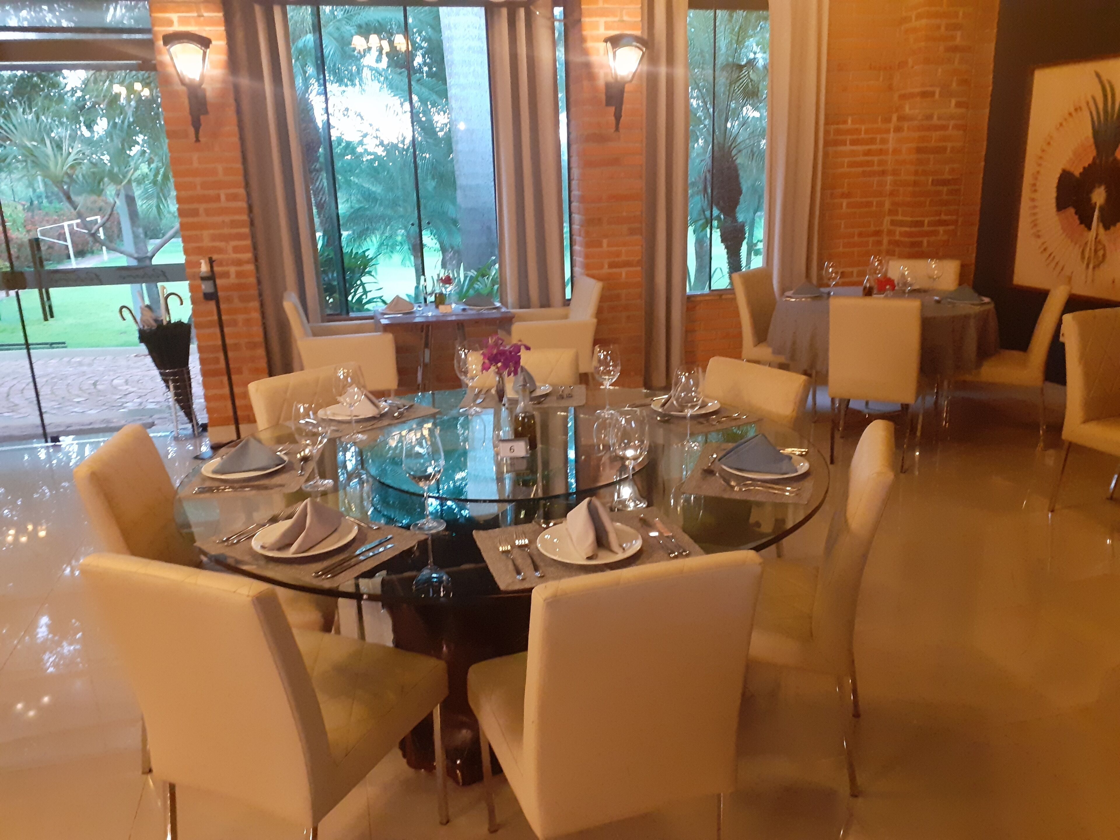 A dining area with a round glass table, white chairs, and a view of greenery through large windows.