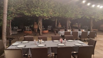 An outdoor dining area with tables set for a meal, surrounded by trees and a stone floor.