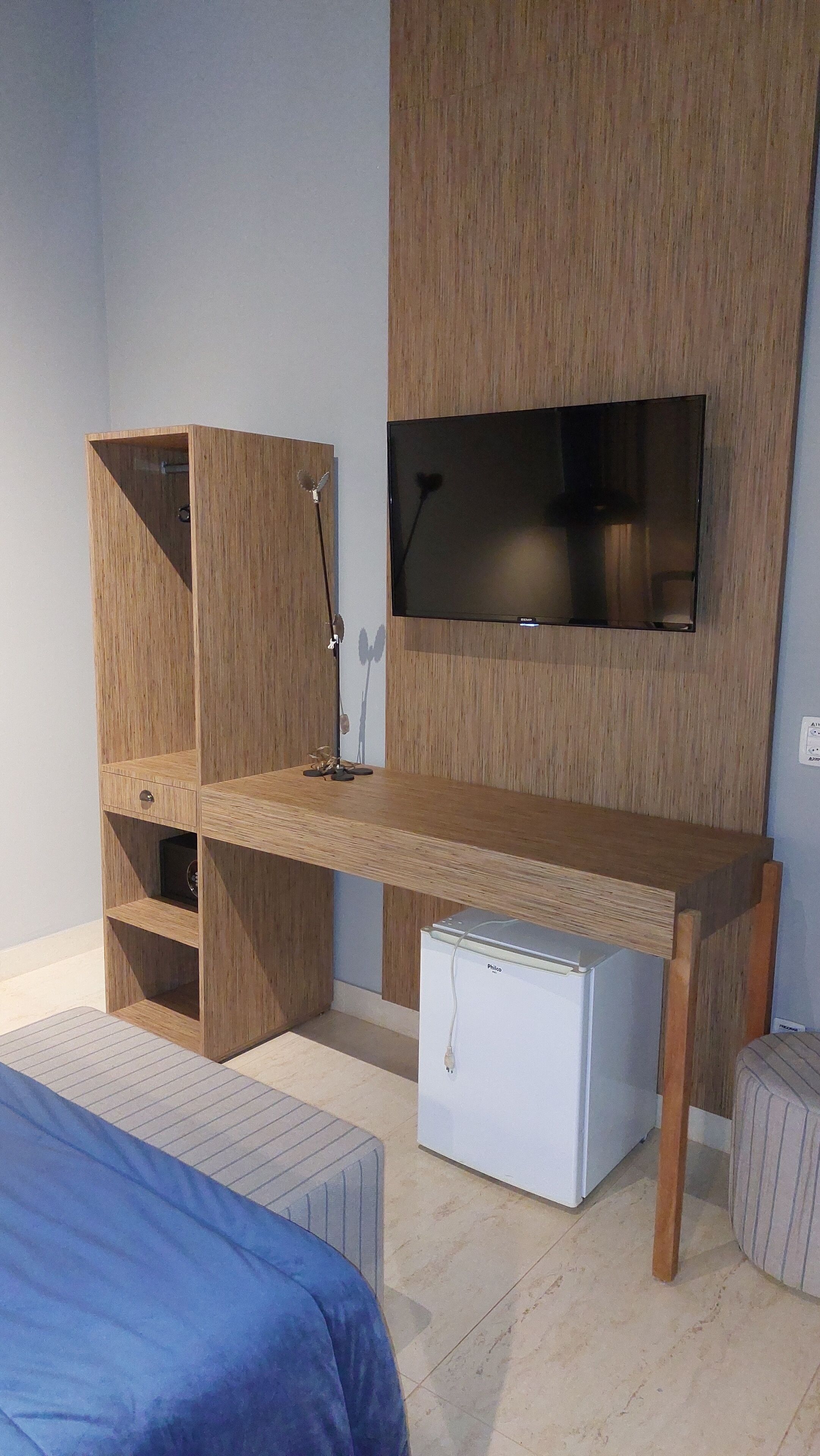 Un dormitorio compacto con un escritorio de madera, un refrigerador blanco pequeño, un televisor montado en la pared y una cama con cubrecama azul.