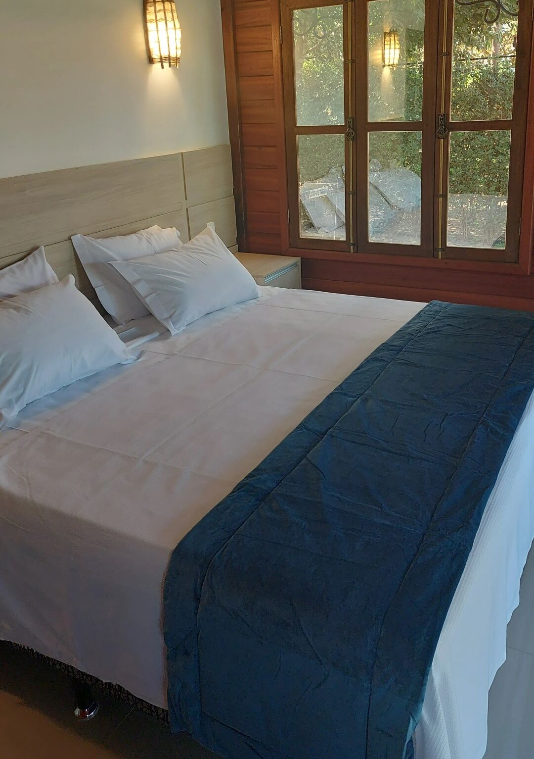Ein Bett mit weißer Bettwäsche und einer blauen Decke, ein Holz-Kopfteil und ein Fenster mit Blick auf Grünflächen.