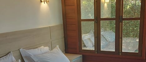 A bed with white bedding and a blue blanket, a wooden headboard, and a window with a view of greenery.