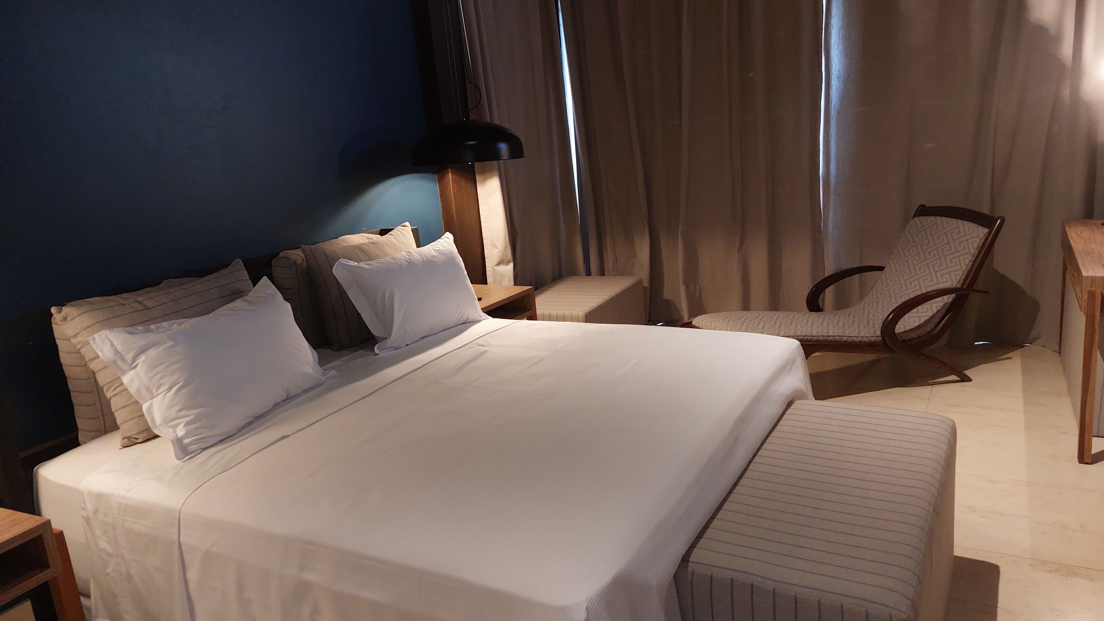 A neatly made bed with white linens and pillows, a wooden nightstand, a chair, and a desk with a lamp.