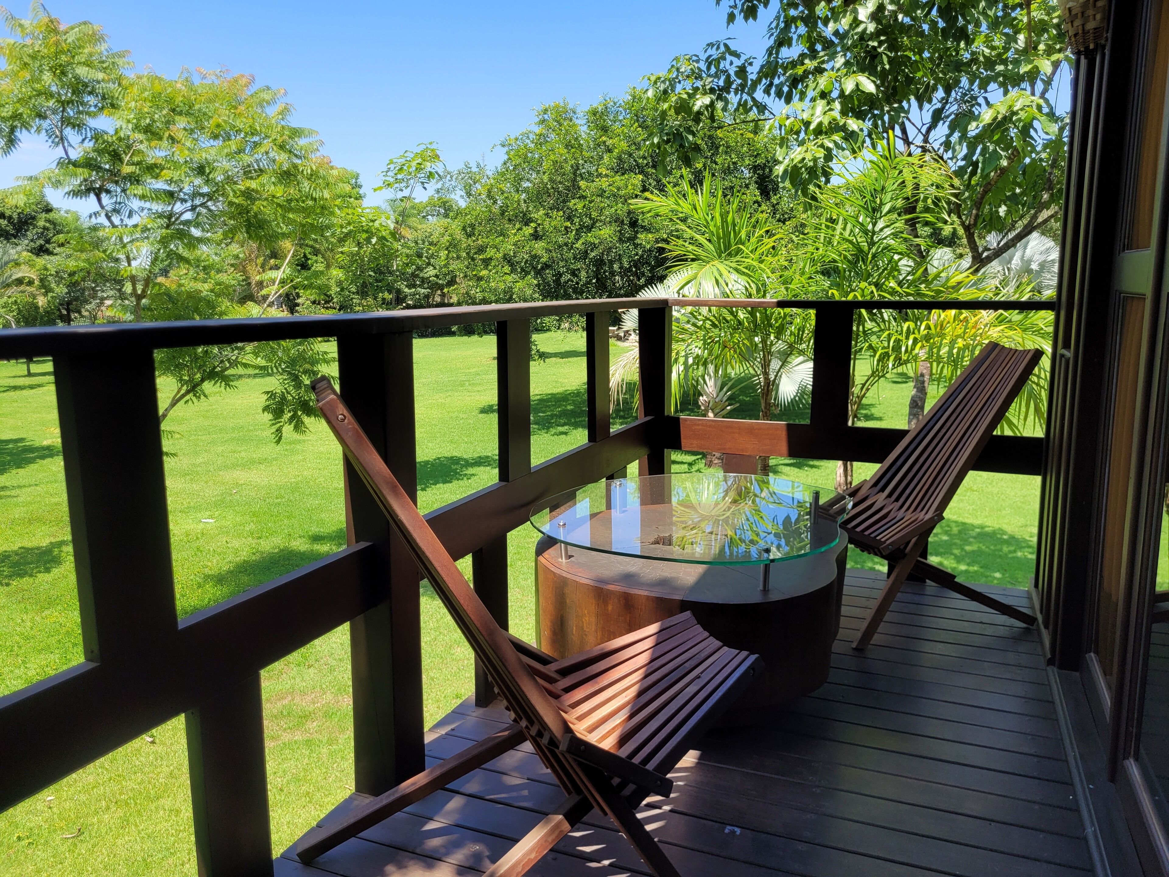 Um deck de madeira com mesa de vidro e duas cadeiras de madeira, com vista para um gramado bem cuidado e árvores.