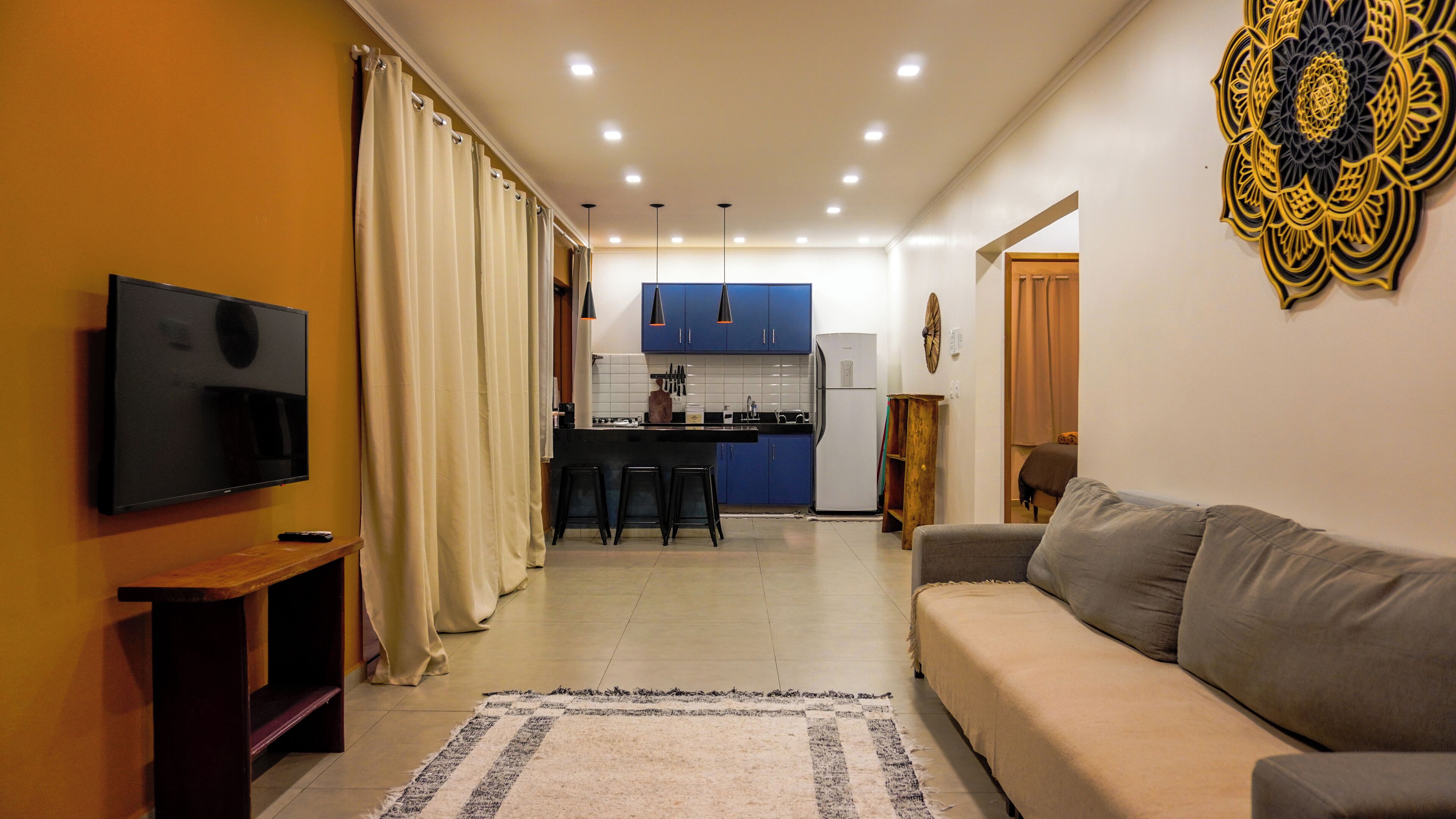 Ein modernes Wohnzimmer mit Flachbildfernseher, grauem Sofa und Küchenzeile mit blauen Schränken und einem weißen Kühlschrank.