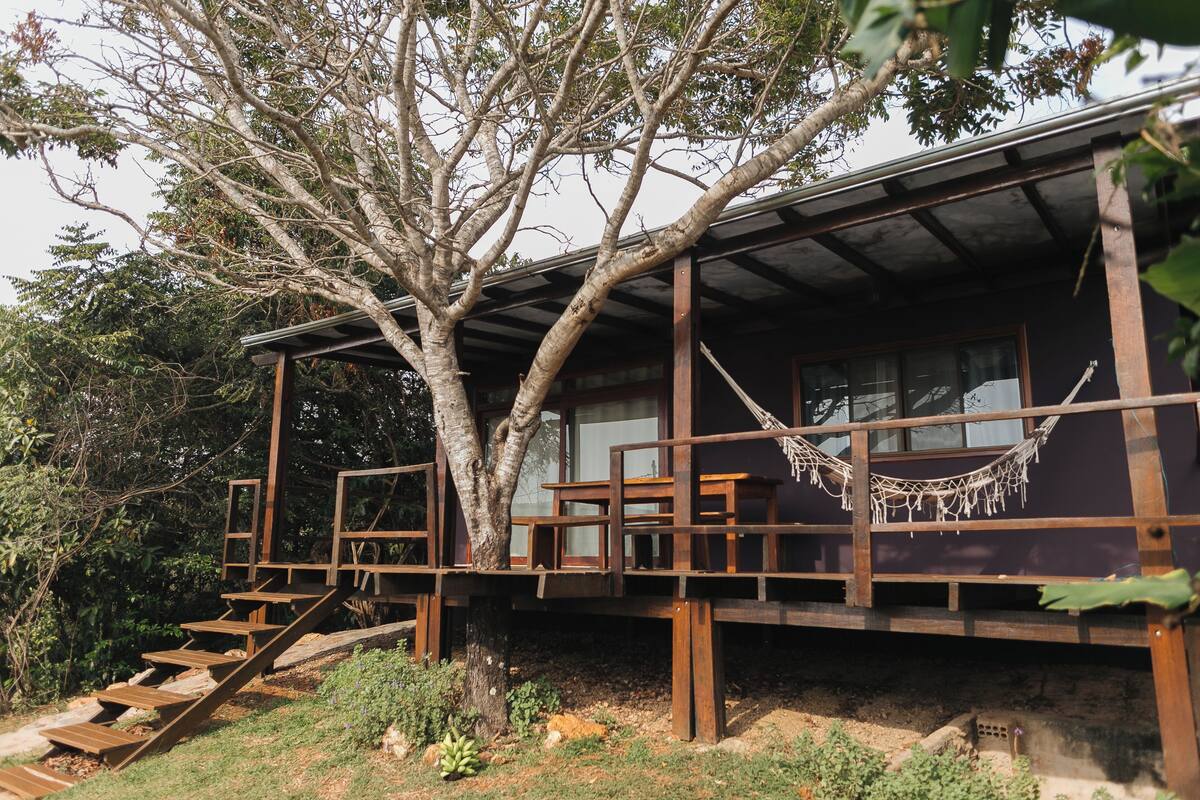 A wooden cabin with a treehouse deck, built-in benches, and a hammock.
