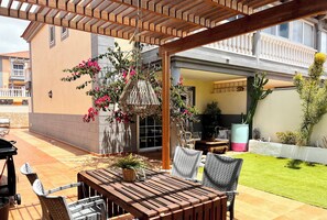 Eine Terrasse im Freien mit Holztisch und Korbstühlen, umgeben von Topfpflanzen und einer hängenden Lampenschirm aus Weidwerk.