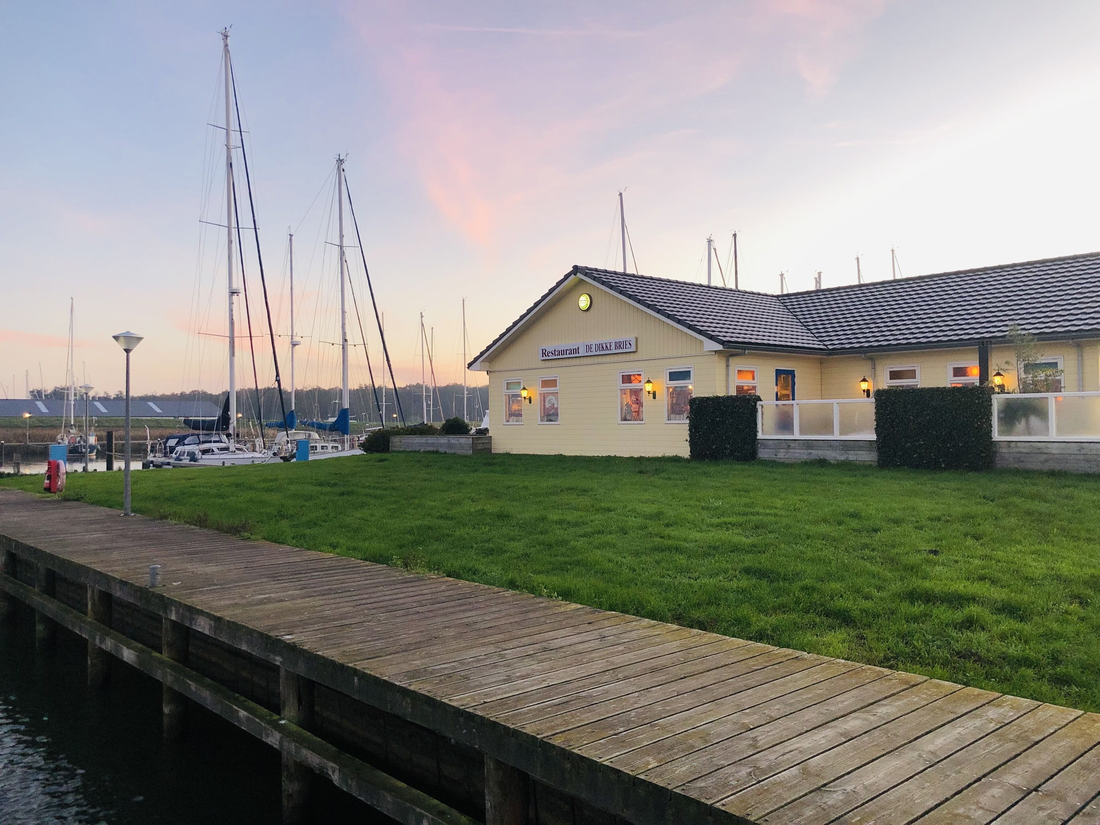 Un restaurant situé au bord de l’eau, avec un quai, des bateaux et une zone herbeuse.