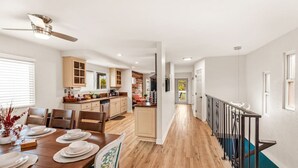 Una cocina amplia con armarios de madera, un comedor con una mesa de madera dispuesta para cuatro personas, y una escalera con barandilla negra que sube a un nivel superior.