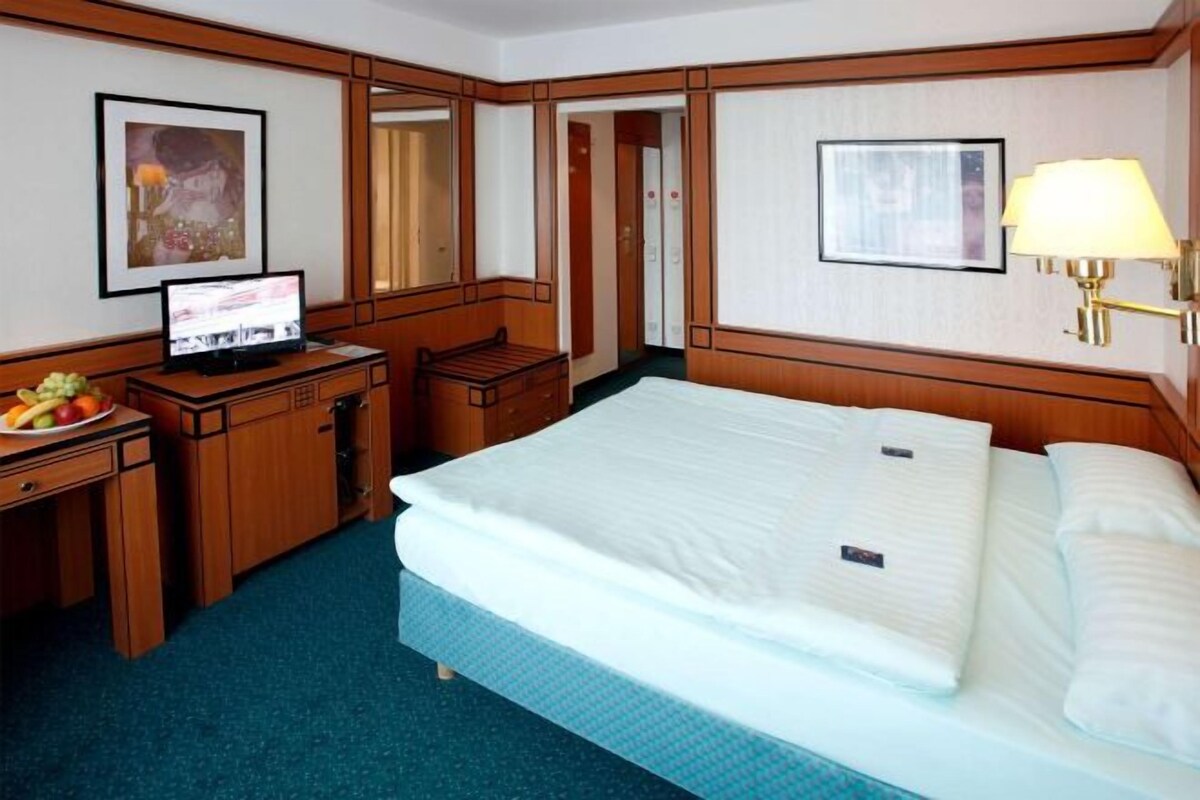 Una habitación de hotel con una cama, un escritorio con computadora, una lámpara y un espejo.