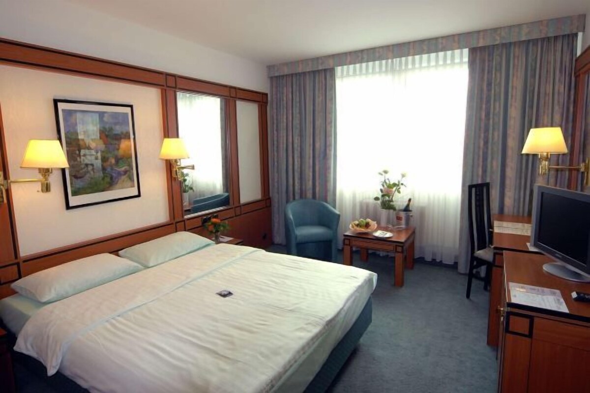 A hotel room with a large bed, two bedside lamps, a desk with a chair, a television, and a window with curtains.
