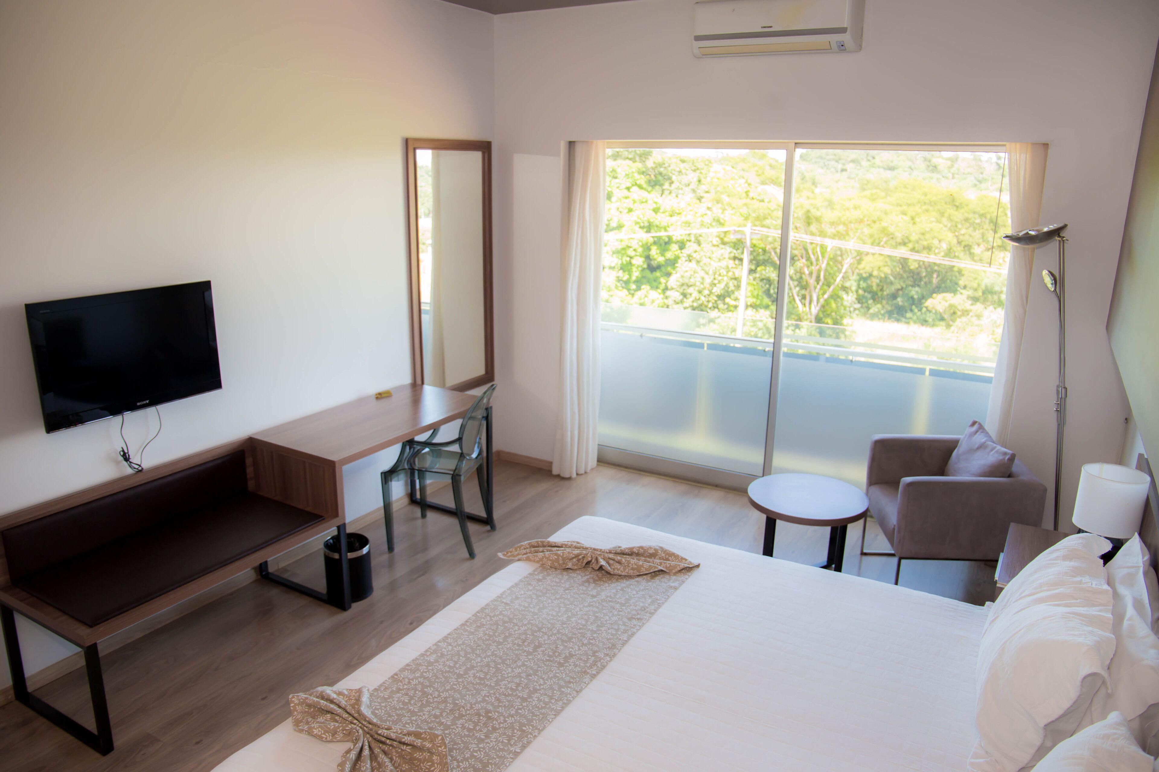 A hotel room with a bed, a desk, a chair, a television, a window with a view of greenery, and a lamp.