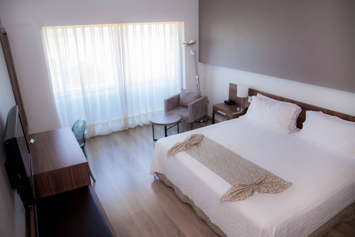 A hotel room with a large bed, a desk, a chair, a lamp, and a window with sheer curtains.