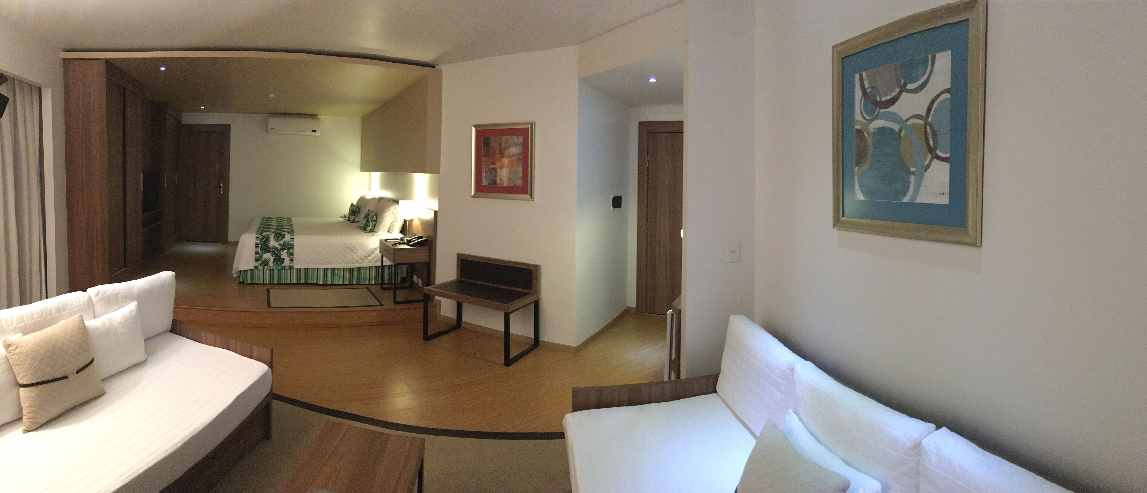 Una camera d'albergo con un letto, un divano, una scrivania e una sedia. C'è un quadro appeso al muro e una porta che dà su un'altra stanza.