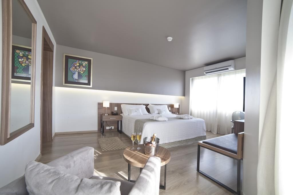 Habitación de hotel con cama, mesitas de noche, una silla, una mesita pequeña con vasos y una botella, y una obra de arte en la pared.