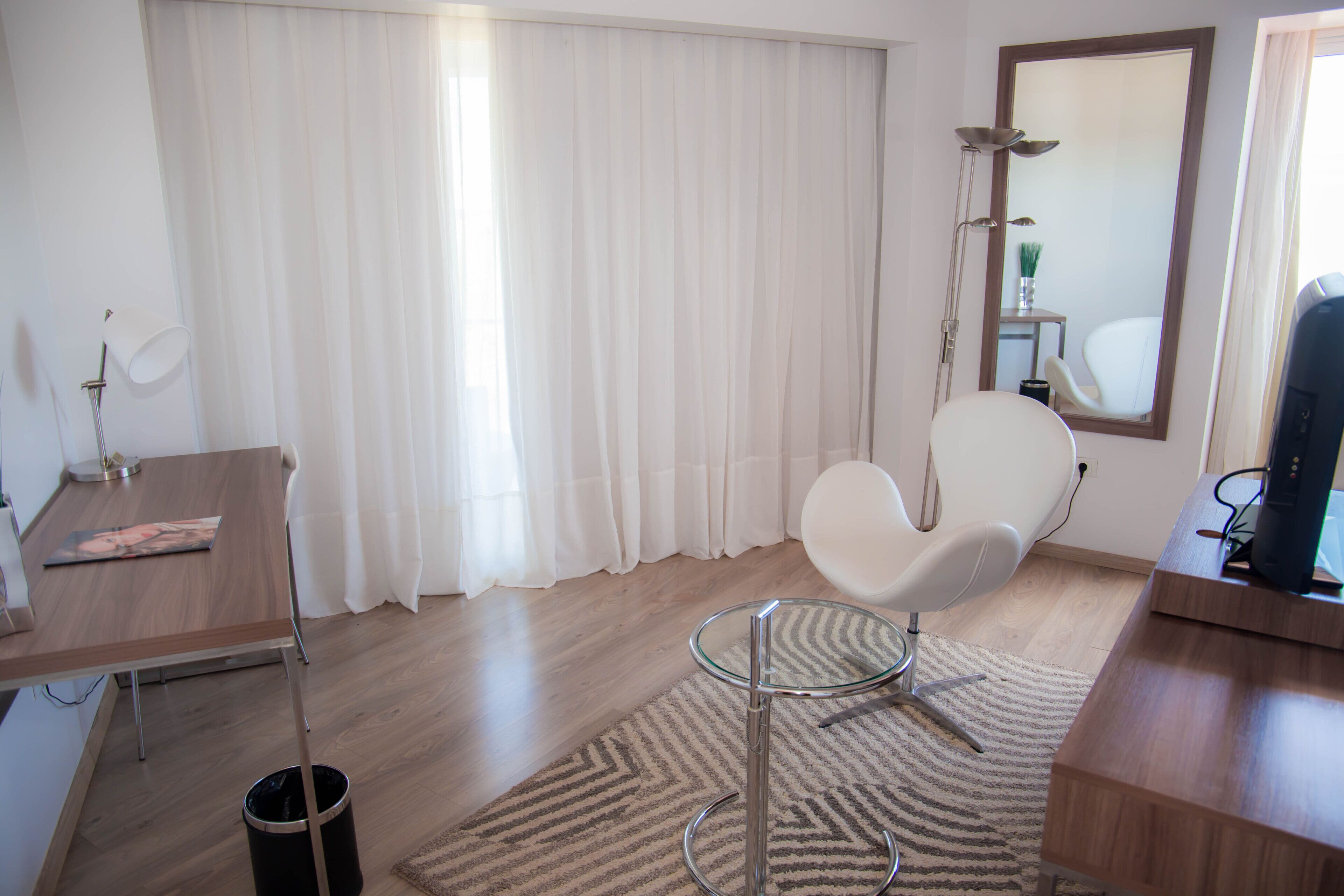 A modern room with a wooden desk, a white chair, a glass table, a large mirror, and a shower area.