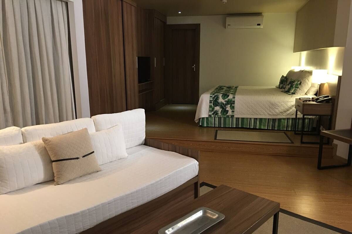 A hotel room with a sofa, a bed with a green and white striped bedspread, a bedside table with a lamp, and a wooden floor.