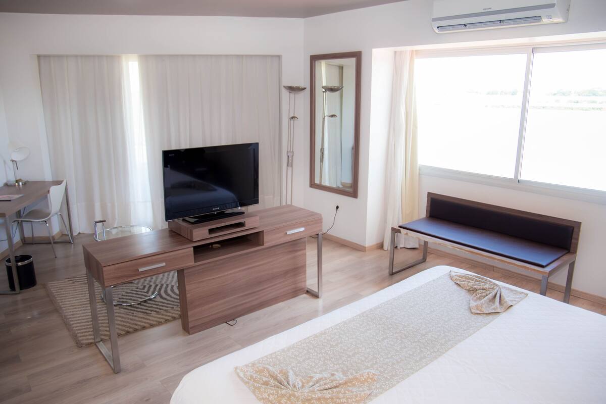 A hotel room with a bed, a desk, a television, a bench, and a large window.