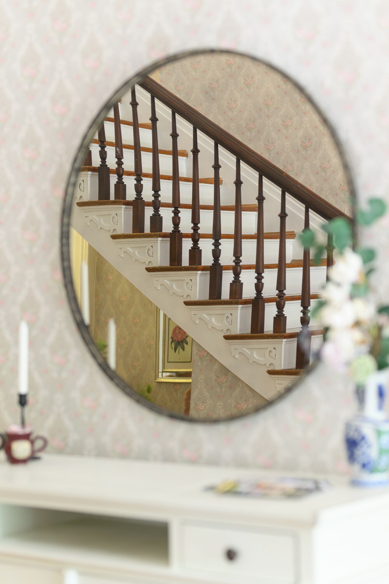 Espejo redondo que refleja una escalera con barandilla de madera y una pared color crema con estampado floral.