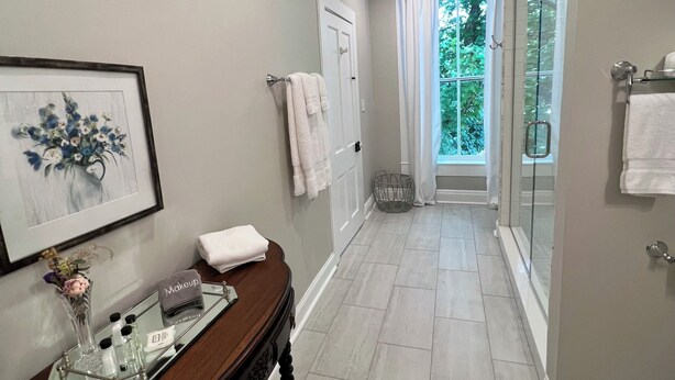 A hallway with a wooden console table, a framed floral artwork, and a glass shower enclosure with a view of greenery. - Rose Hill Inn (Versailles)