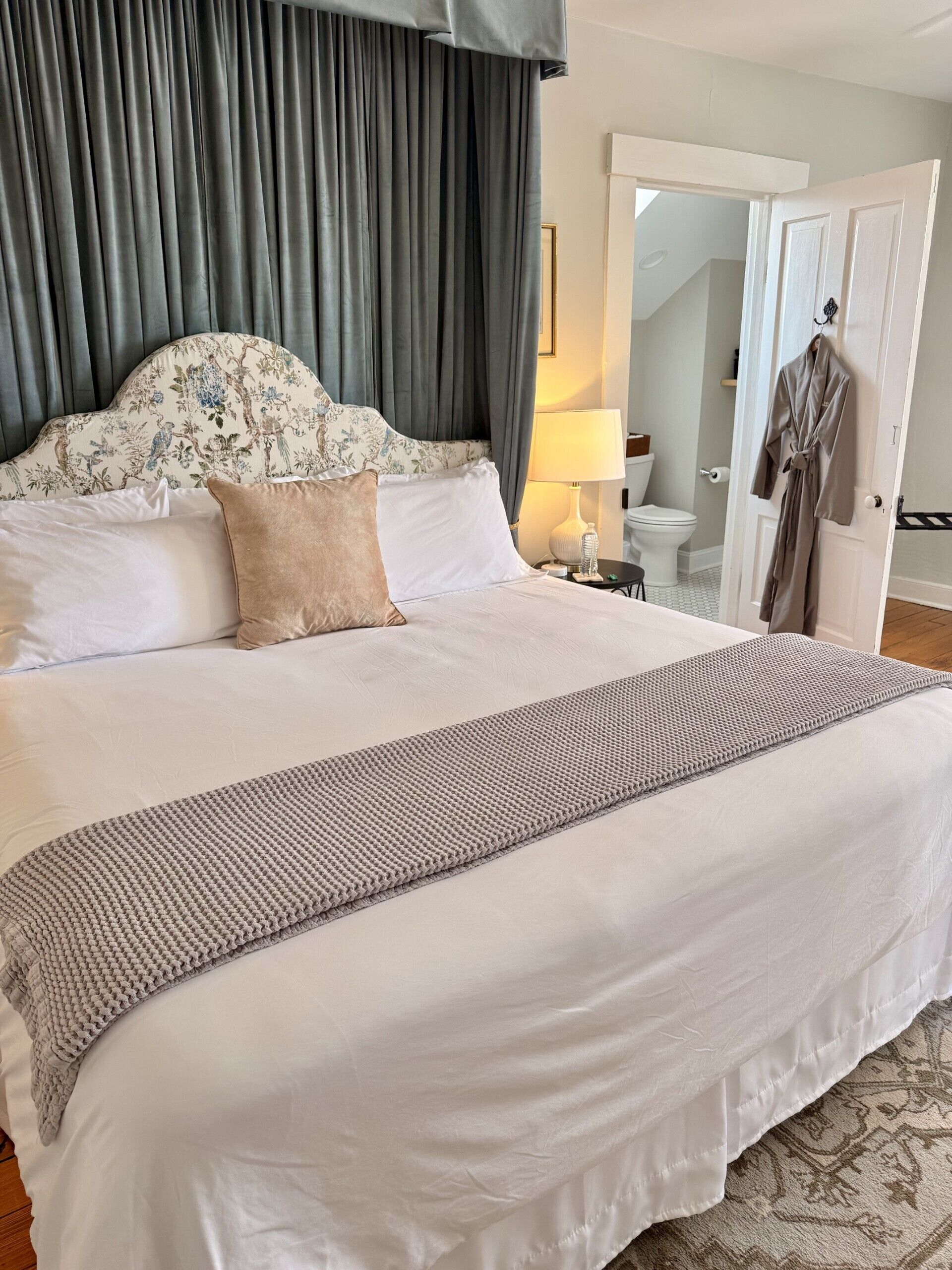 Cama bem arrumada com cabeceira estampada, abajur e roupão pendurado na porta.