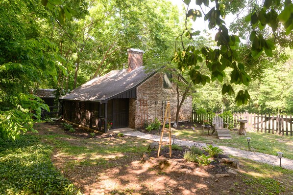 A rustic stone building with a sloped roof, surrounded by lush greenery and a wooden fence. - Rose Hill Inn (Versailles)