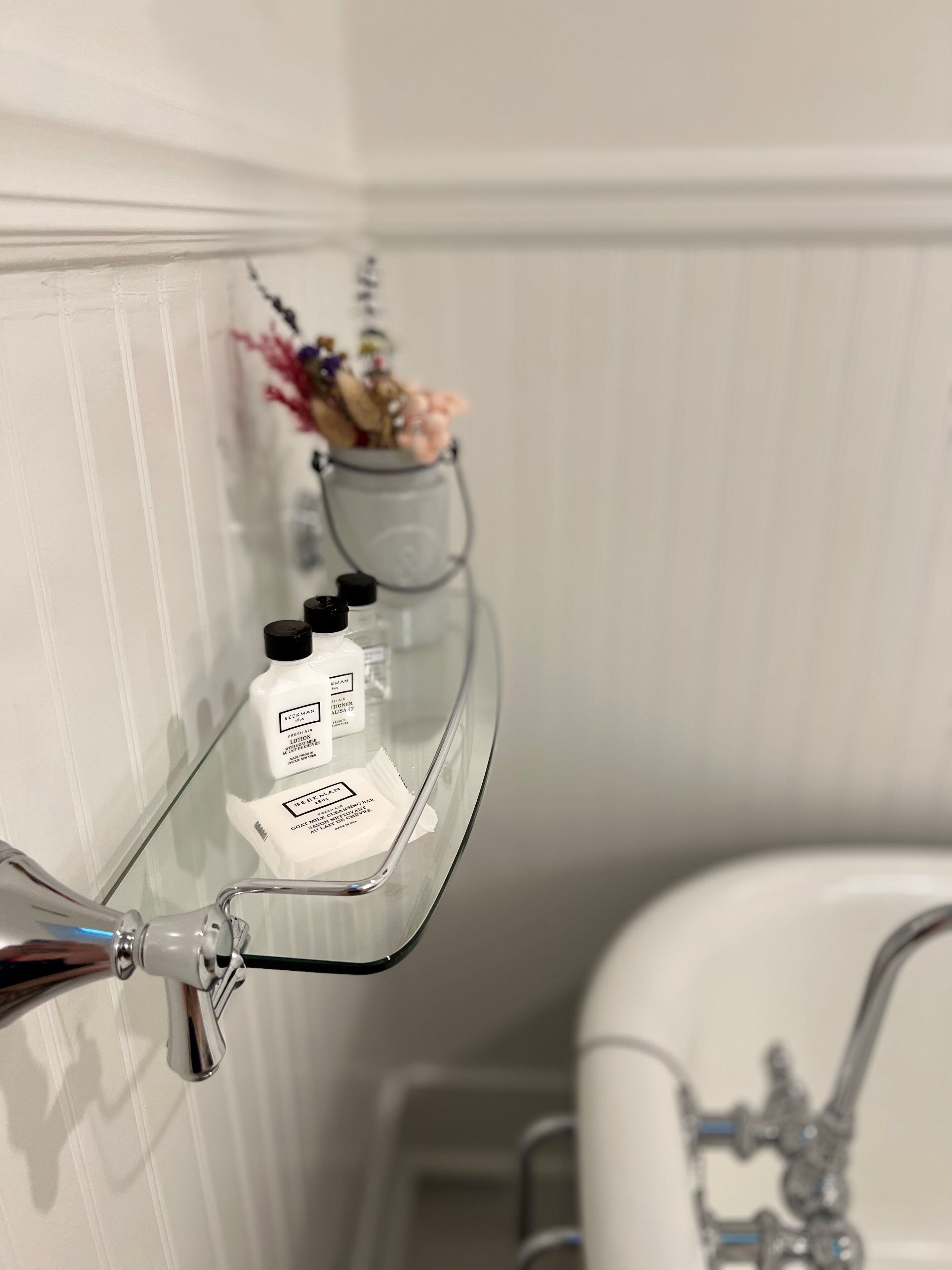 Une salle de bain avec une étagère en verre où sont rangés les articles de toilette, une baignoire blanche et une petite plante en arrière-plan.