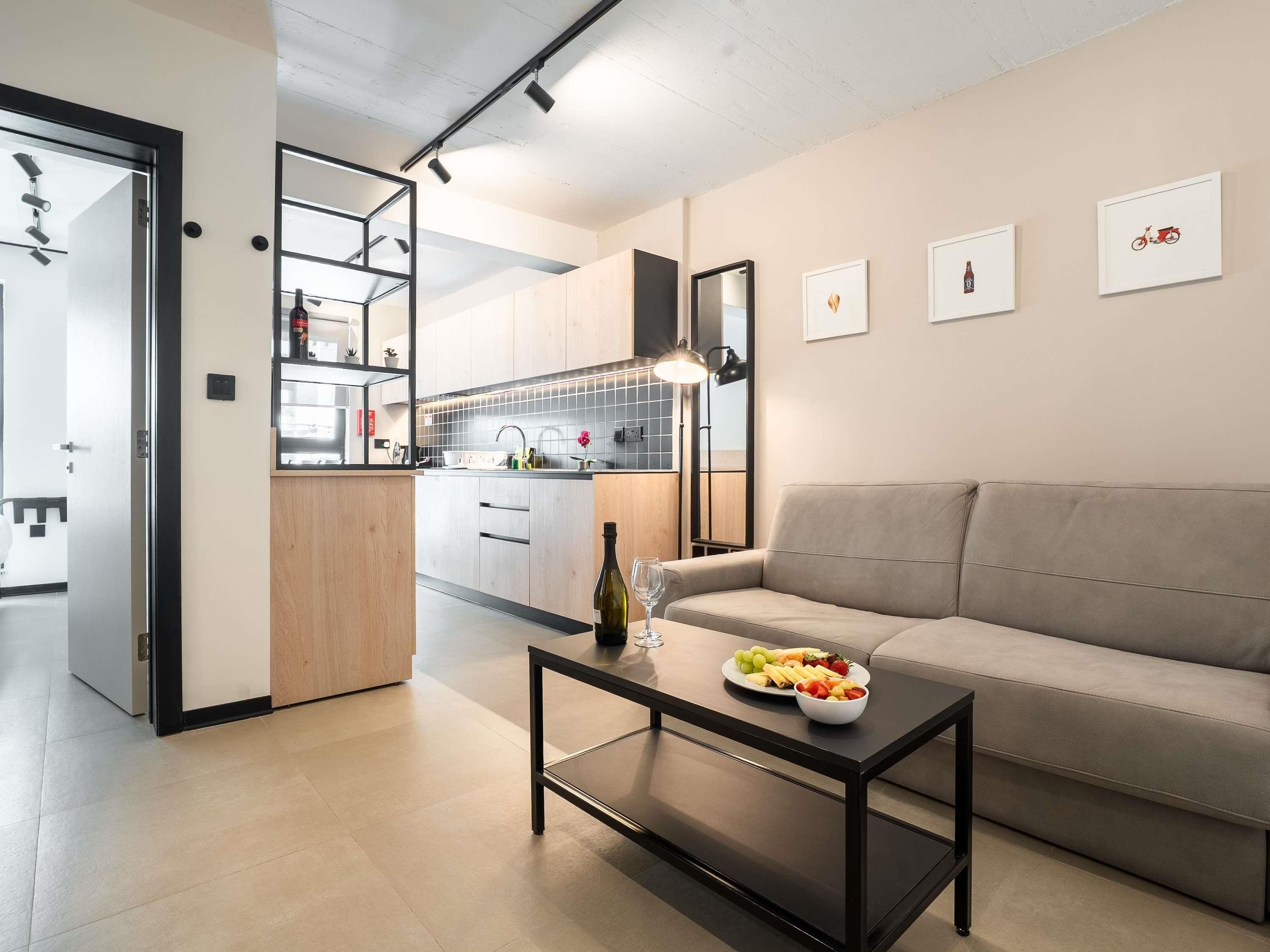 Una sala de estar moderna con un sofá, una mesa baja con fruta, y una cocina al fondo.