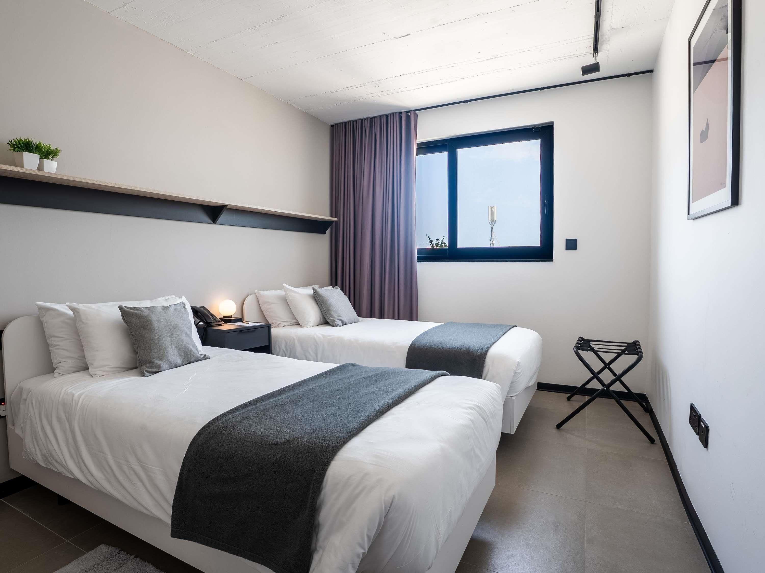 A hotel room with two beds, a window, a nightstand, a lamp, a small table, and a wall-mounted shelf.