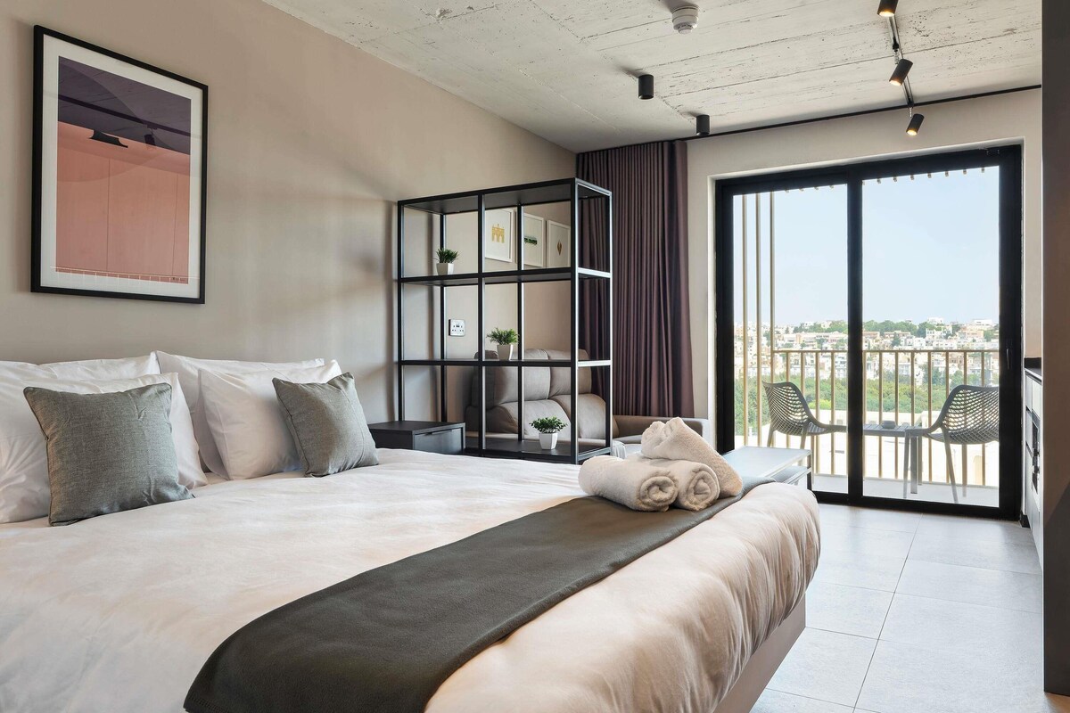 A bedroom with a large bed, a built-in bookshelf, a balcony with chairs, and a city view.