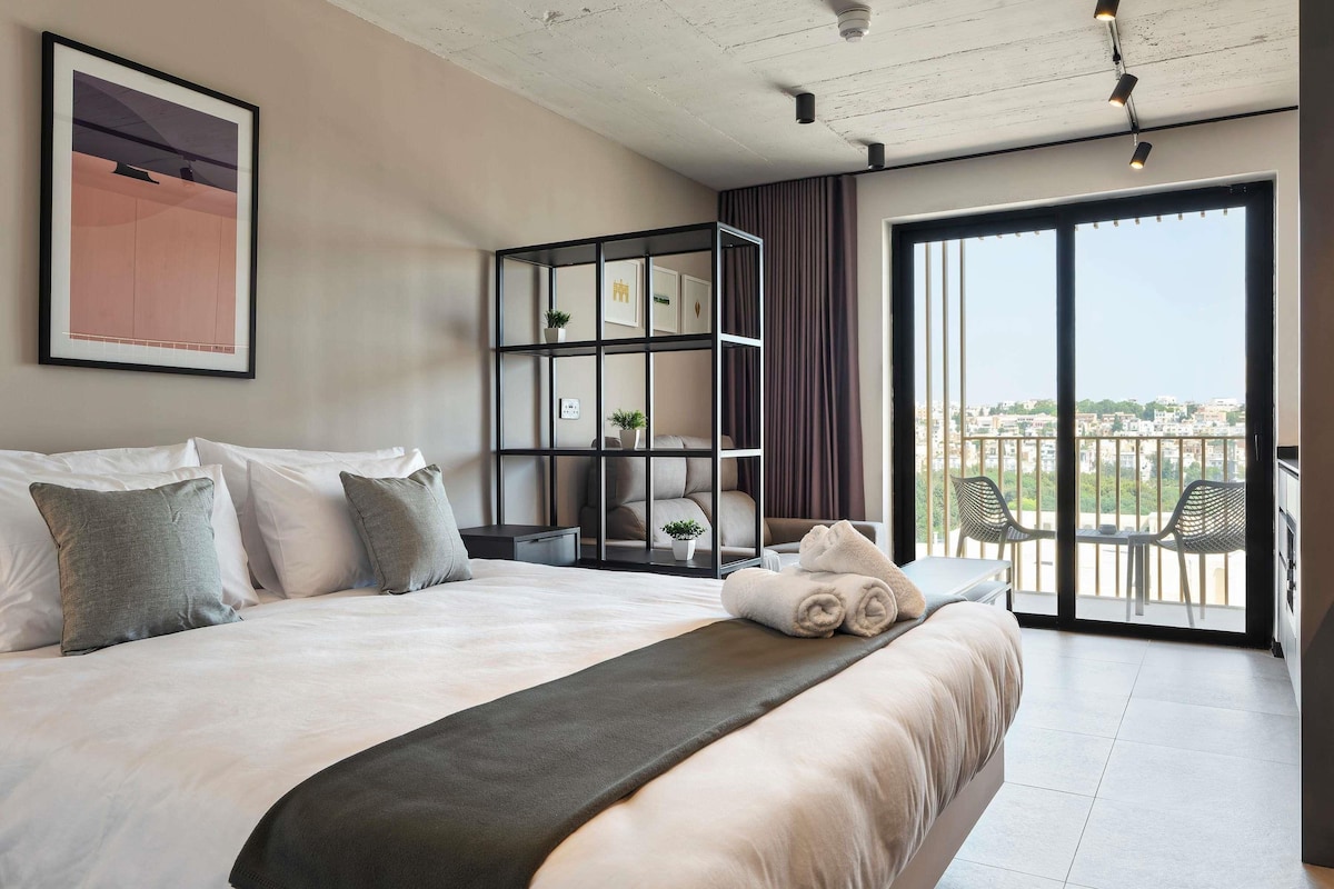 A bedroom with a large bed, a built-in bookshelf, a balcony with chairs, and a city view.