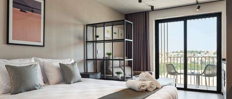 A bedroom with a large bed, a built-in bookshelf, a balcony with chairs, and a city view.