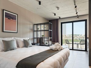 A bedroom with a large bed, a built-in bookshelf, a balcony with chairs, and a city view. - Aparthotel Adagio Malta Central (Msida)