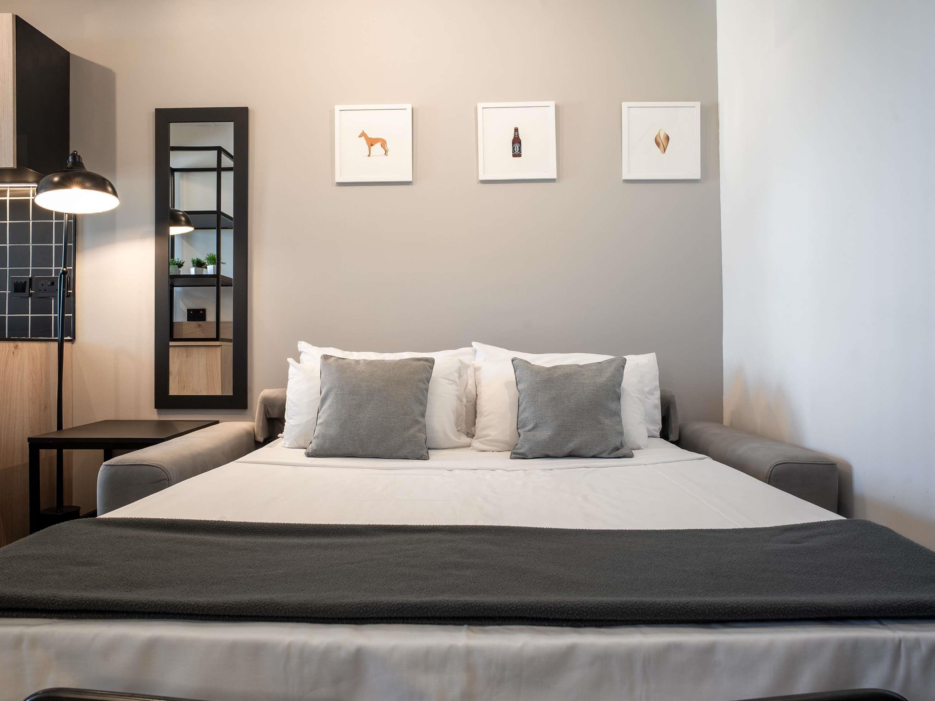 A modern bedroom with a large bed, grey pillows, a dark throw blanket, and three framed artworks on the wall.